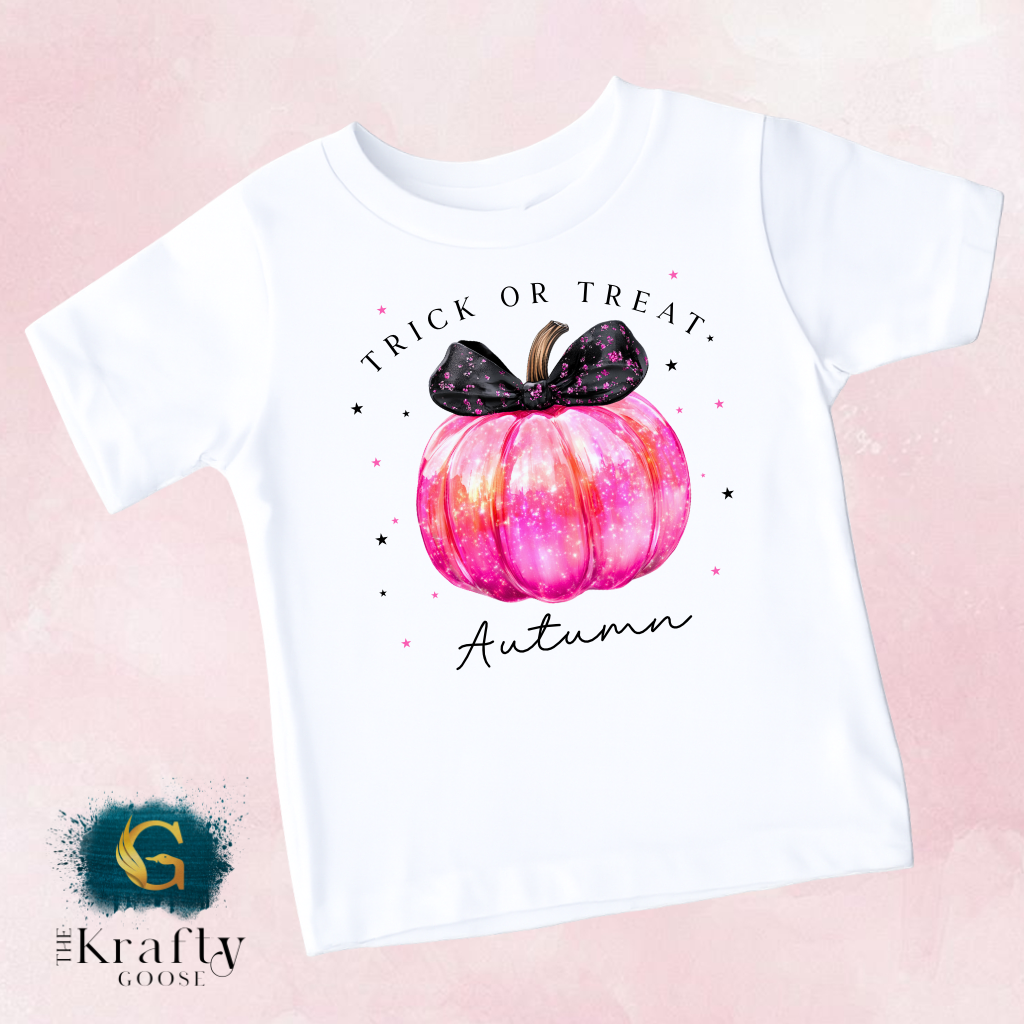 A white t-shirt with a graphic design featuring a pink pumpkin with a black bow and the text "Trick or Treat Autumn" printed on it.