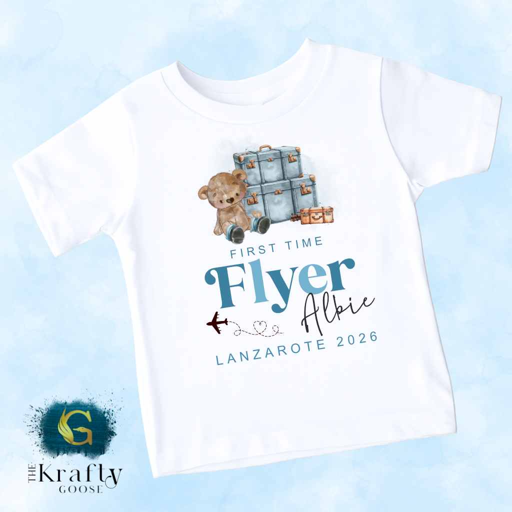 A white t-shirt with a graphic design featuring a teddy bear and suitcases, along with text that says "First Time Flyer Albie Lanzarate 2026".