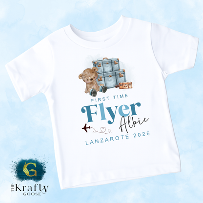 A white t-shirt with a graphic design featuring a teddy bear and suitcases, along with text that says "First Time Flyer Albie Lanzarate 2026".