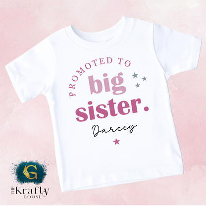 A white t-shirt with text that reads "PROMOTED TO BIG SISTER. Darcey" printed on it.
