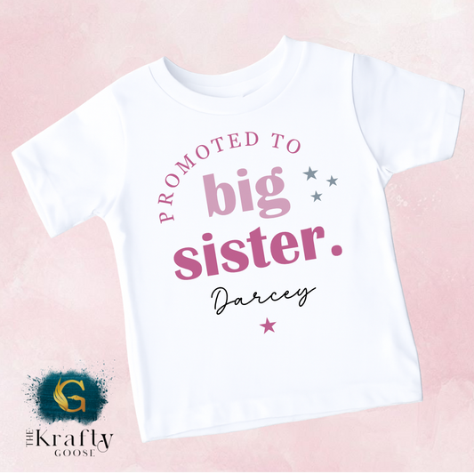 A white t-shirt with text that reads "PROMOTED TO BIG SISTER. Darcey" printed on it.