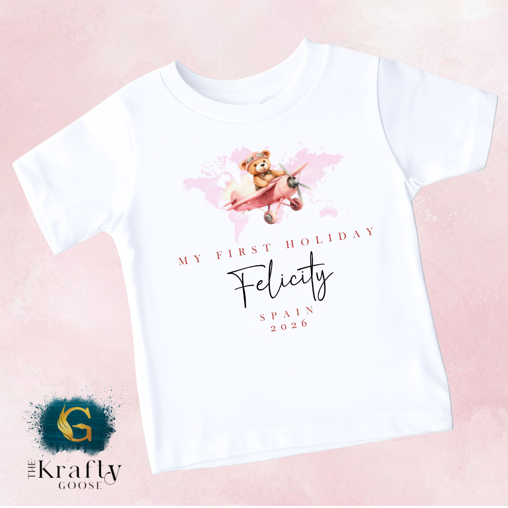 The image is a white t-shirt with a graphic design featuring a teddy bear flying in a pink airplane. The design includes text that reads "My first holiday" and "Felicity Spain 2026".