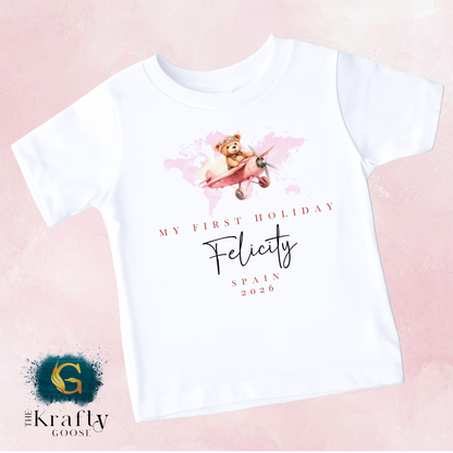 The image is a white t-shirt with a graphic design featuring a teddy bear flying in a pink airplane. The design includes text that reads "My first holiday" and "Felicity Spain 2026".