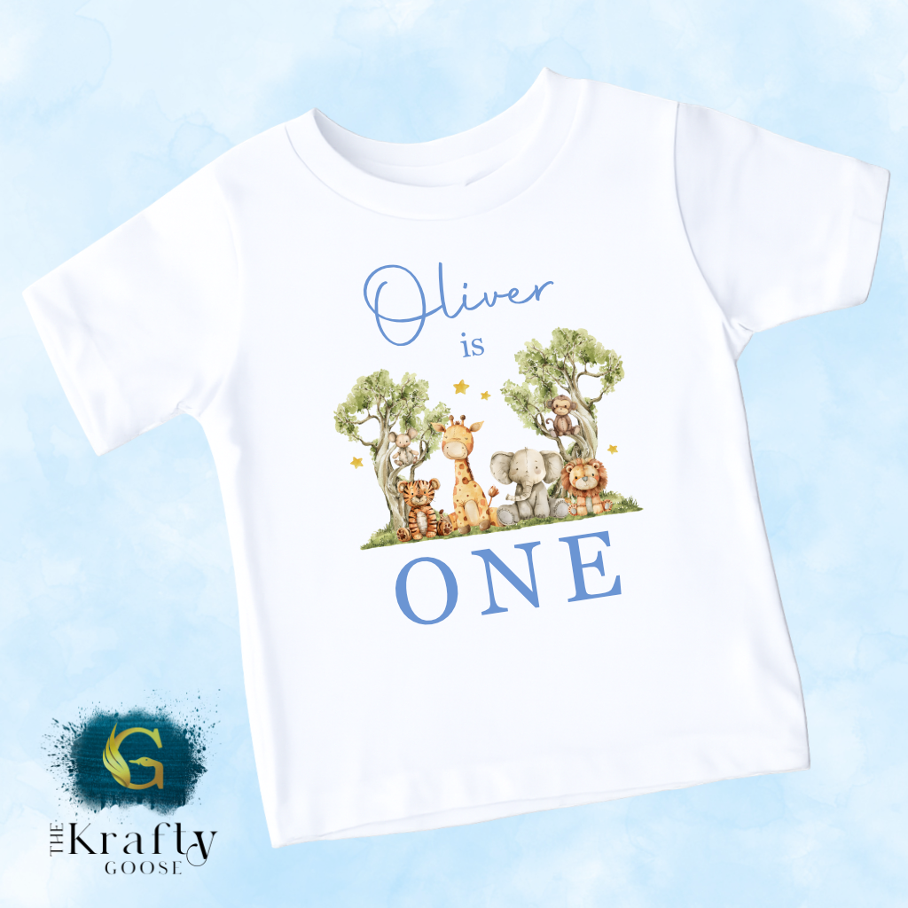 A white t-shirt with a graphic design featuring a giraffe, elephant, and other animals, along with the text "Oliver is ONE" and a blue background.