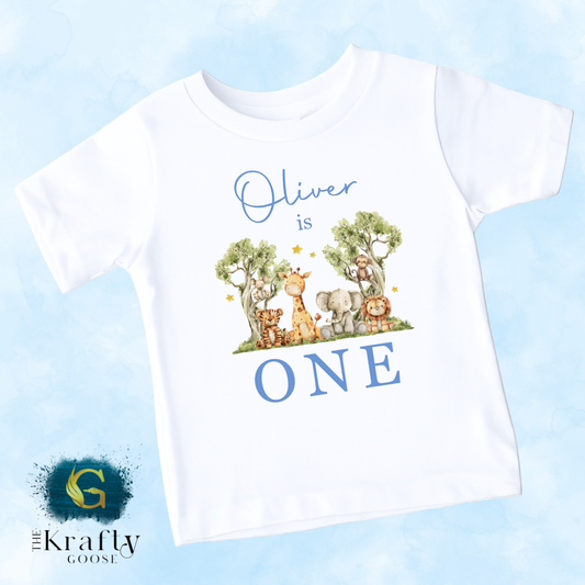A white t-shirt with a graphic design featuring a giraffe, elephant, and other animals, along with the text "Oliver is ONE" and a blue background.
