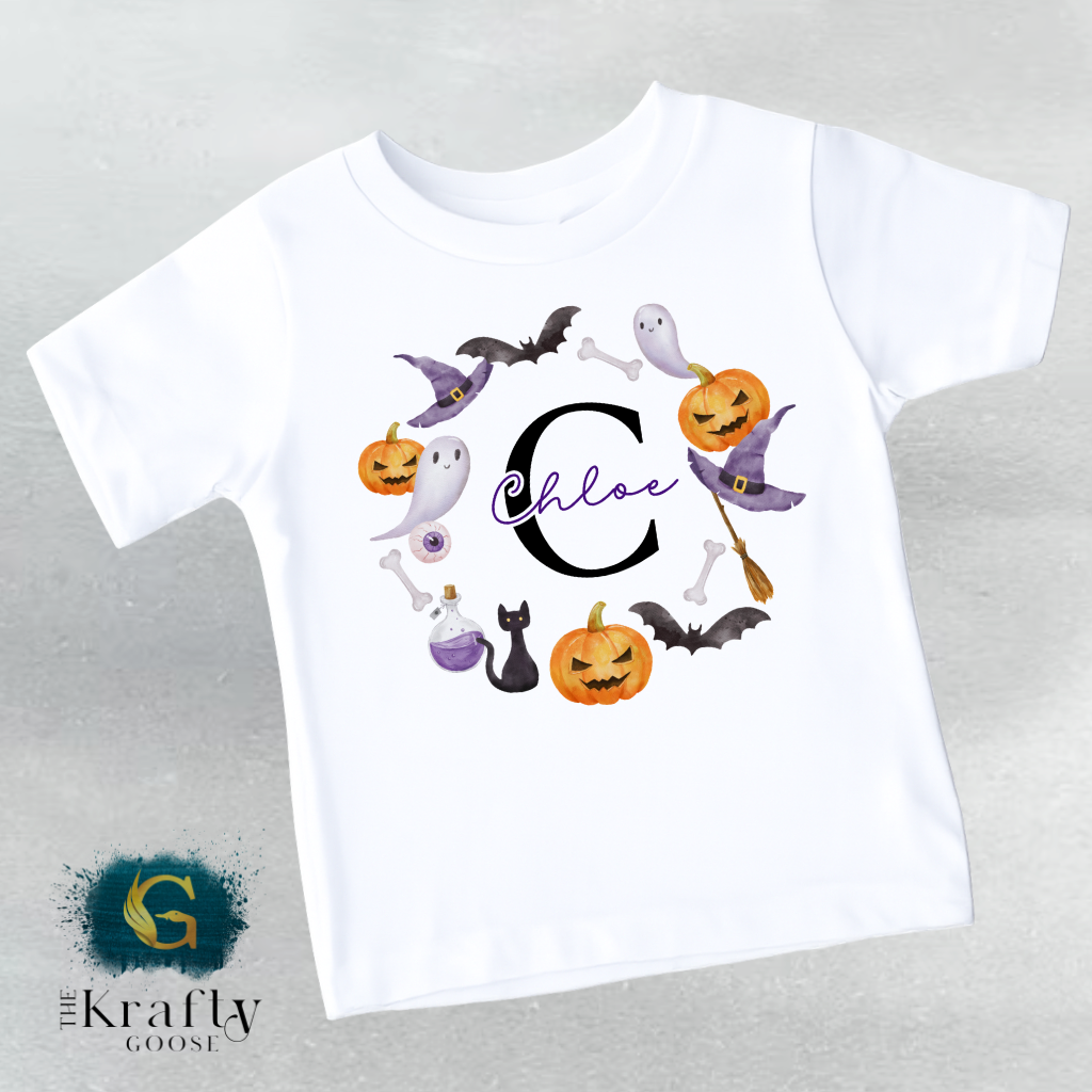 A white t-shirt with a Halloween-themed design featuring a large letter "C" surrounded by various spooky and festive elements such as bats, pumpkins, and a cat.