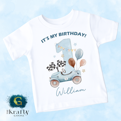 A white t-shirt with a graphic design featuring a car, balloons, and a number one, along with the text "It's my birthday!" and the name "William" written below.