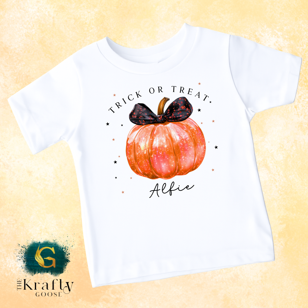 A white t-shirt with a graphic design featuring a pumpkin and a black bow, along with the text "Trick or Treat" and "Alfie" printed on it.