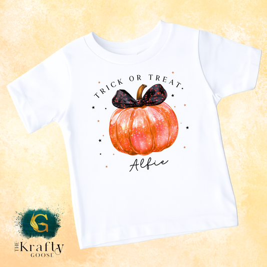 A white t-shirt with a graphic design featuring a pumpkin and a black bow, along with the text "Trick or Treat" and "Alfie" printed on it.