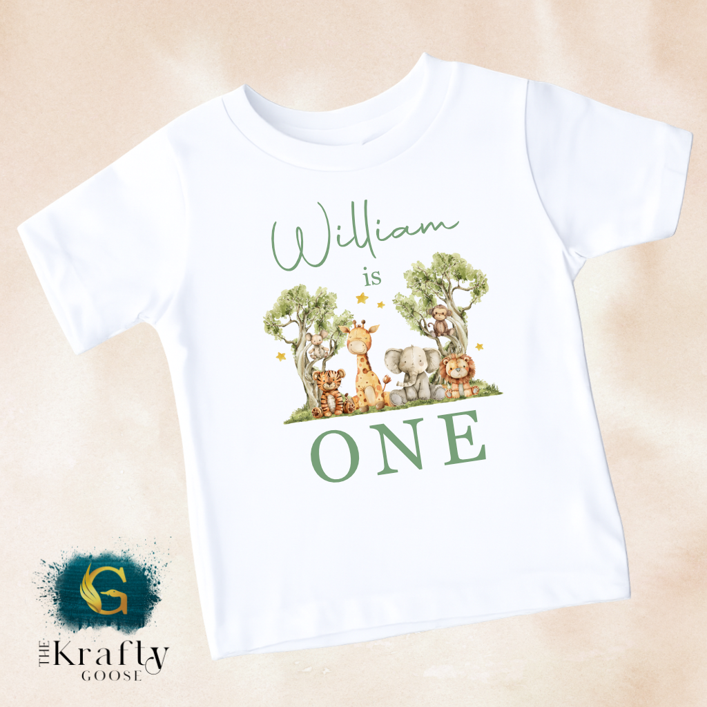 A white t-shirt with a graphic design featuring a group of animals, including a giraffe, lion, and elephant, along with the text "William is ONE" printed on it.