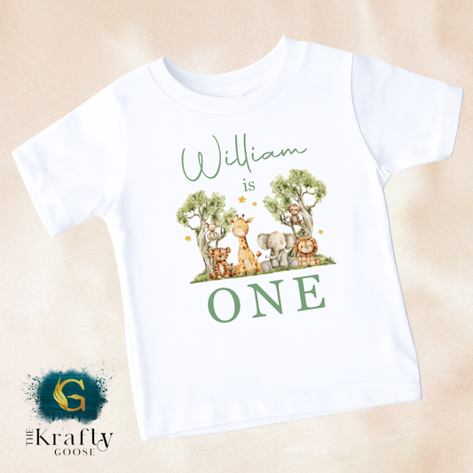 A white t-shirt with a graphic design featuring a group of animals, including a giraffe, lion, and elephant, along with the text "William is ONE" printed on it.