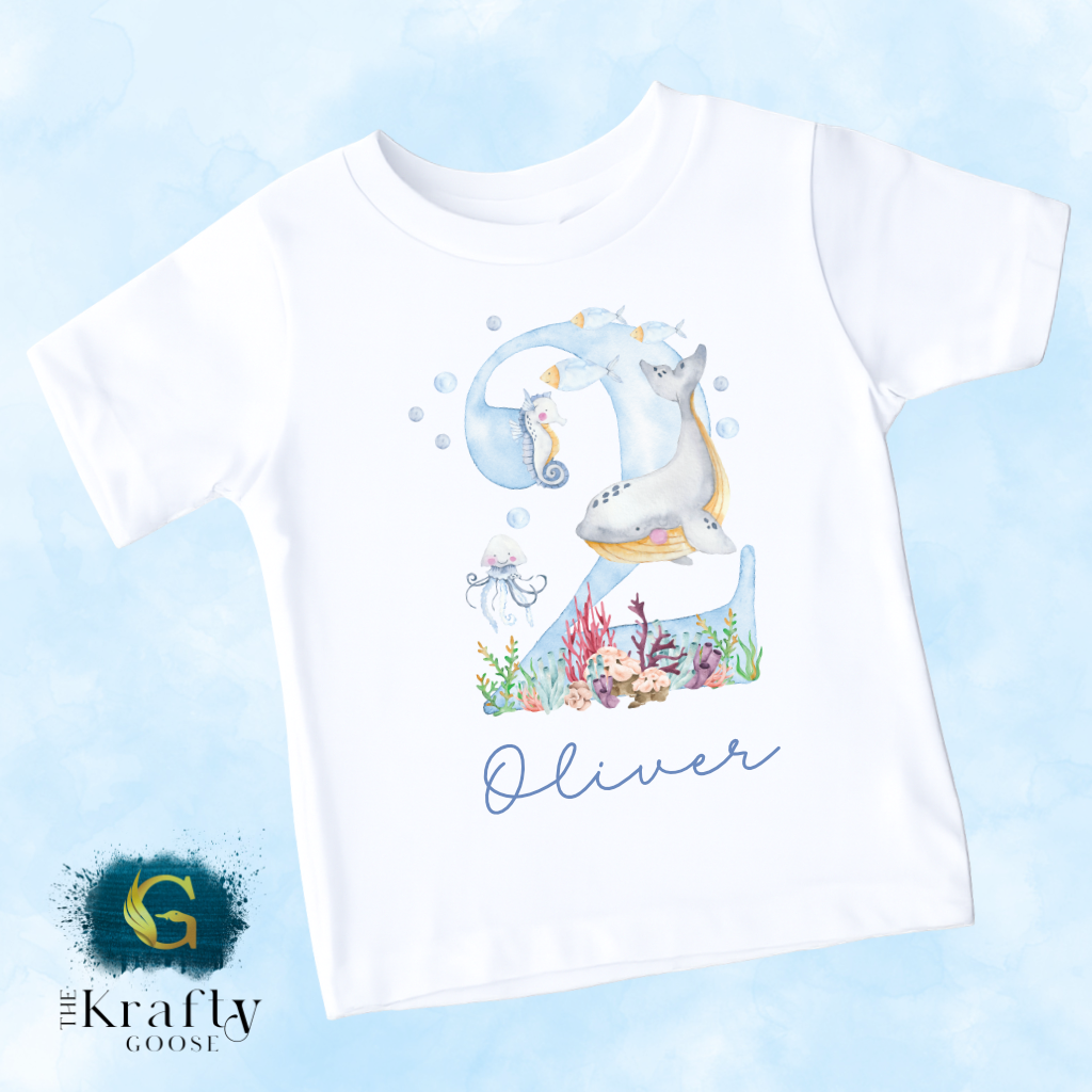 A white t-shirt with a watercolor illustration of a seahorse and other marine life, along with the name "Oliver" written on it.