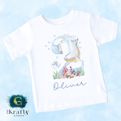 A white t-shirt with a watercolor illustration of a seahorse and other marine life, along with the name "Oliver" written on it.