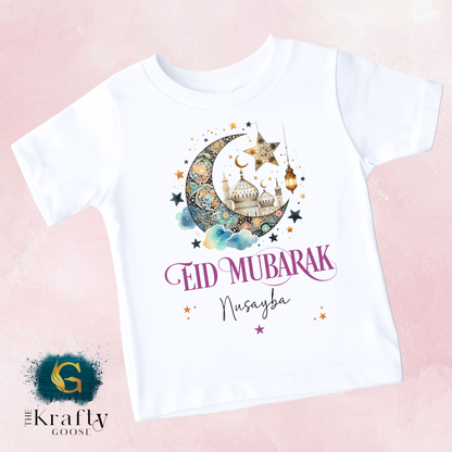 A white t-shirt with a design featuring a crescent moon, star, and mosque, along with the text "Eid Mubarak Nusayba" printed on it.