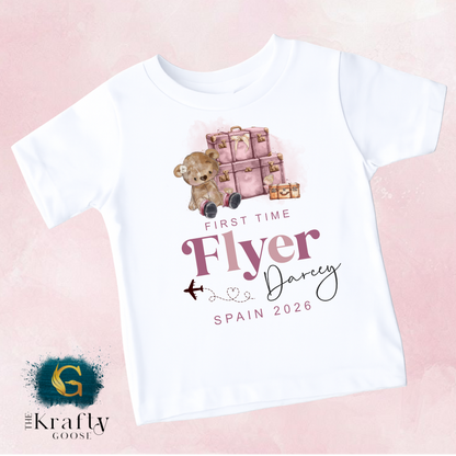 The image is a white t-shirt with a graphic design featuring a teddy bear and a suitcase, along with text that reads "First time flyer Darrey Spain 2026".