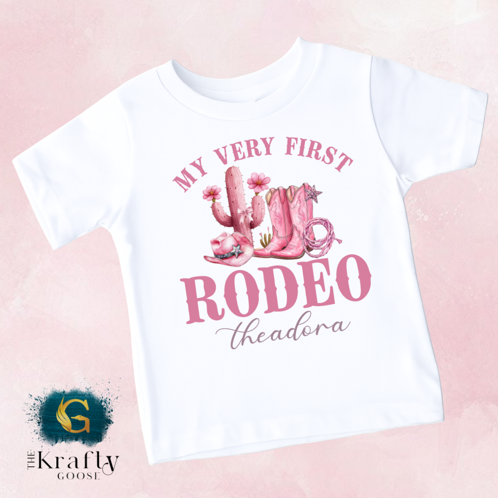 A white t-shirt with a pink graphic design featuring a cactus, cowboy boots, and the text "MY VERY FIRST RODEO THEADORA" printed on it.