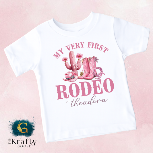 A white t-shirt with a pink graphic design featuring a cactus, cowboy boots, and the text "MY VERY FIRST RODEO THEADORA" printed on it.