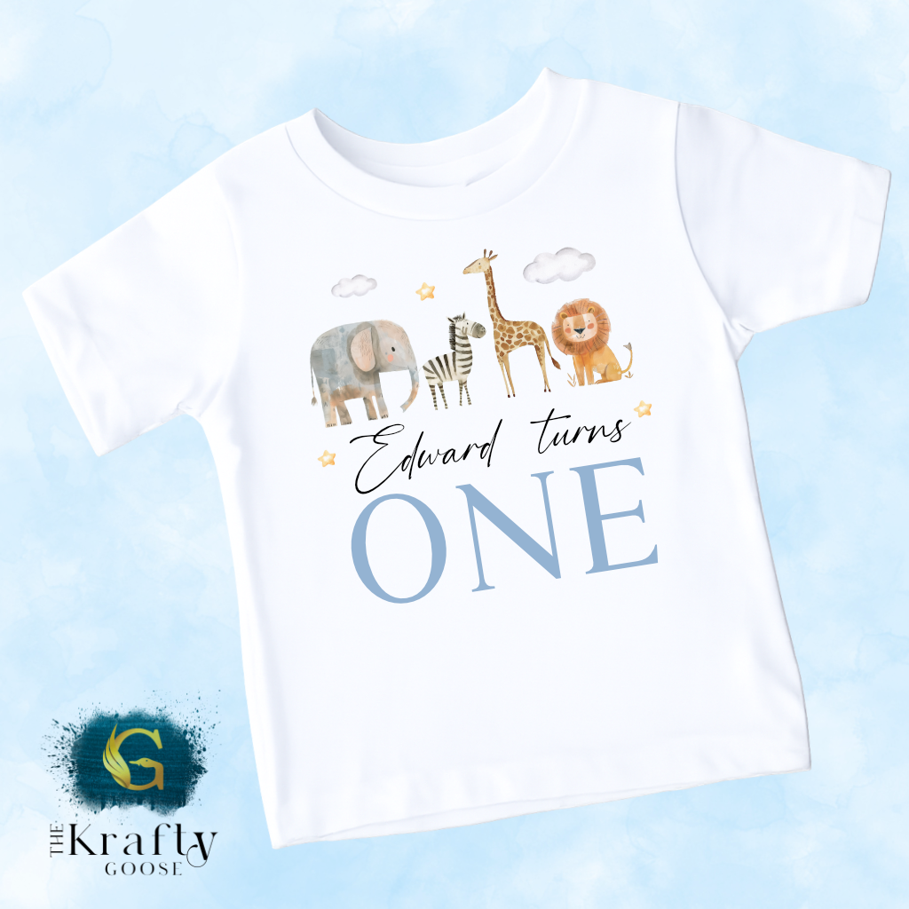 A white t-shirt with a graphic design featuring a giraffe, elephant, and lion, along with the text "Edward turns one".