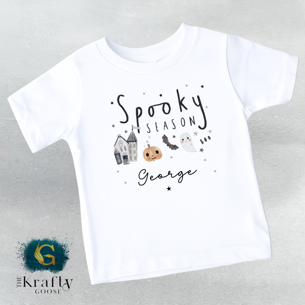 A white t-shirt with a Halloween-themed design, including a ghost, a pumpkin, and a castle. The text on the t-shirt says "Spooky Season George".