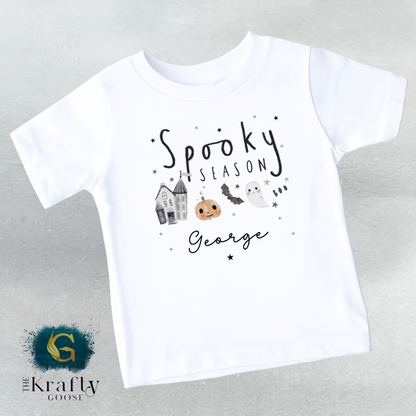 A white t-shirt with a Halloween-themed design, including a ghost, a pumpkin, and a castle. The text on the t-shirt says "Spooky Season George".
