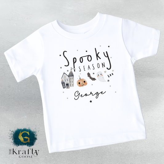 A white t-shirt with a Halloween-themed design, including a ghost, a pumpkin, and a castle. The text on the t-shirt says "Spooky Season George".