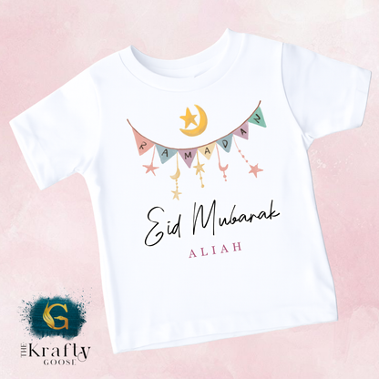 A white t-shirt with a graphic design featuring a crescent moon, stars, and the text "Eid Mubarak Aliah" printed on it.