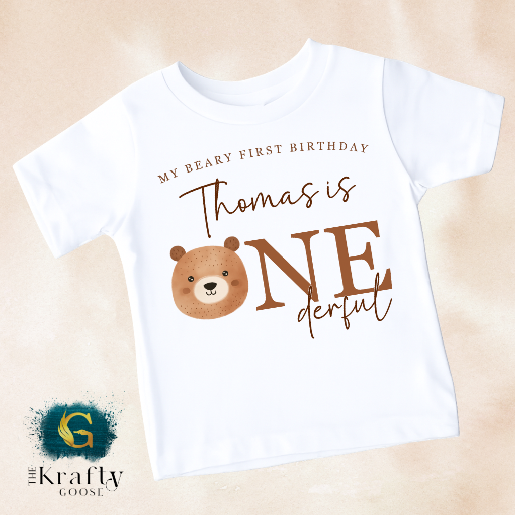 A white t-shirt with a brown bear graphic and text that says "My Beary First Birthday Thomas is One derful" printed on it.