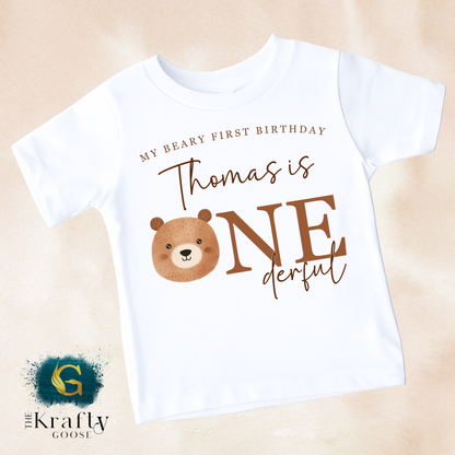 A white t-shirt with a brown bear graphic and text that says "My Beary First Birthday Thomas is One derful" printed on it.