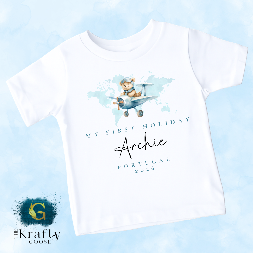 A white t-shirt with a graphic design of a teddy bear flying in a plane, along with text that says "My first holiday Archie Portugal 2026".