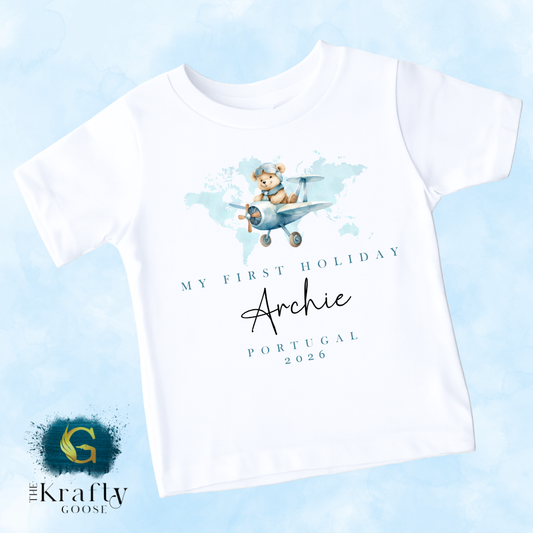 A white t-shirt with a graphic design of a teddy bear flying in a plane, along with text that says "My first holiday Archie Portugal 2026".