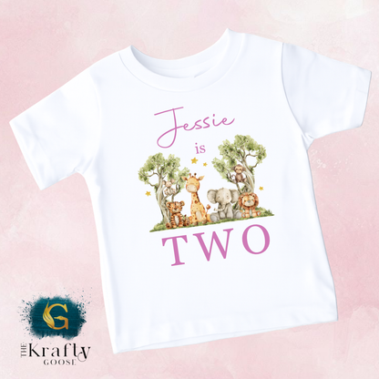A white t-shirt with a graphic design featuring a group of animals, including giraffes, elephants, and lions, along with the text " Jessie is two".