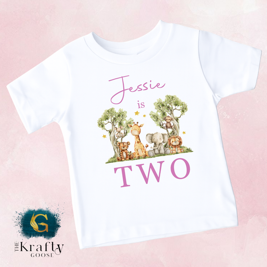A white t-shirt with a graphic design featuring a group of animals, including giraffes, elephants, and lions, along with the text " Jessie is two".