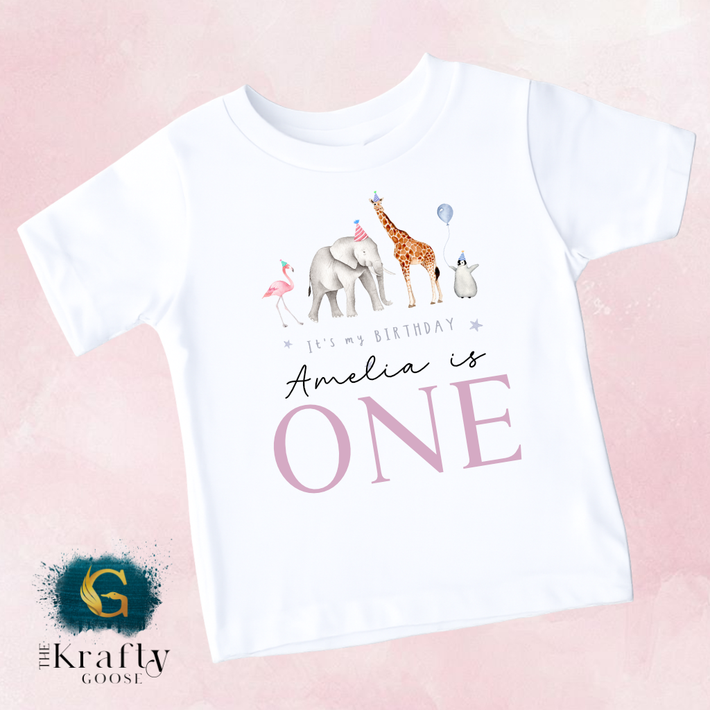 A white t-shirt with a graphic design featuring a giraffe, elephant, and flamingo, along with the text "It's my birthday Amelia is ONE" and the logo for "The Krusty Goose".