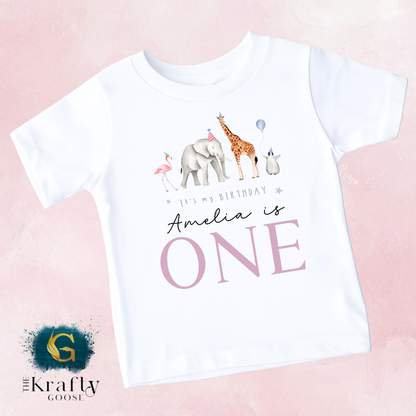 A white t-shirt with a graphic design featuring a giraffe, elephant, and flamingo, along with the text "It's my birthday Amelia is ONE" and the logo for "The Krusty Goose".