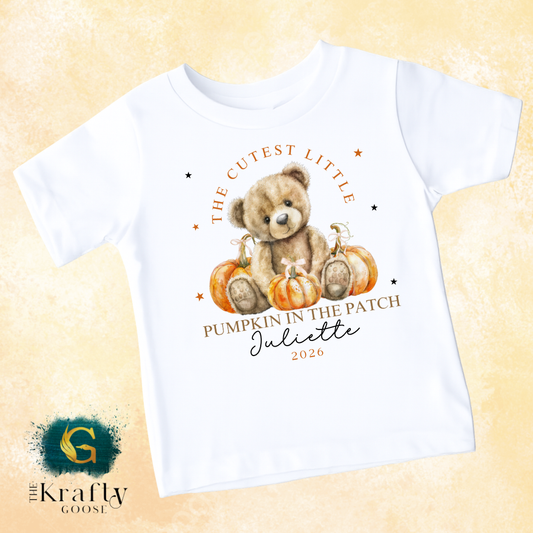 A white t-shirt with a printed design featuring a teddy bear holding a pumpkin and surrounded by stars. The text on the t-shirt reads "The cutest little PUMPKIN IN THE PATCH JULIETTE 2020".